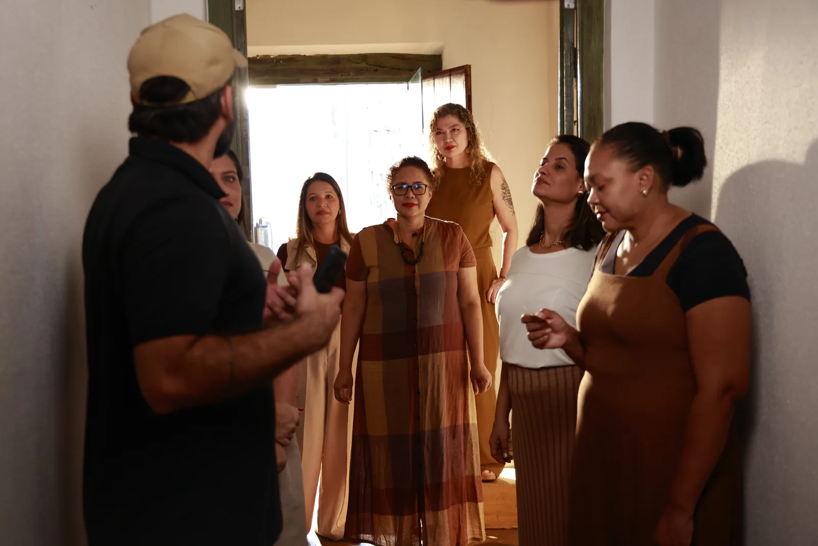 Mulheres reunidas no corredor do casarão, diante de uma porta aberta por onde entra a luz, durante a gravação da campanha.